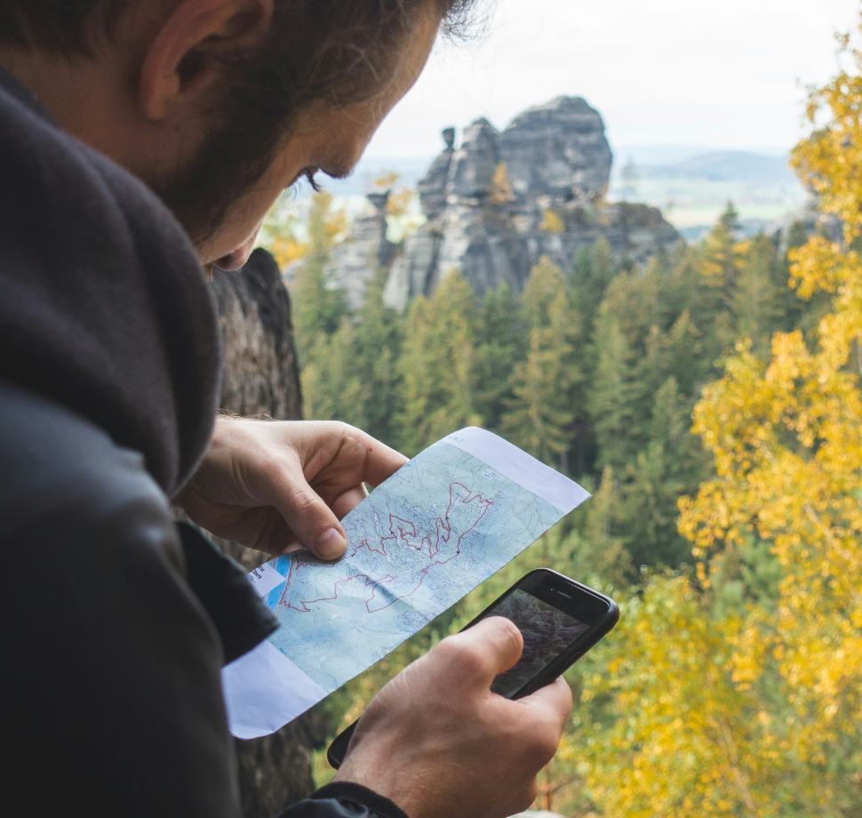 Mann mit Karte und Handy in den Händen. Im Hintergrund ist ein Wald zu sehen. 